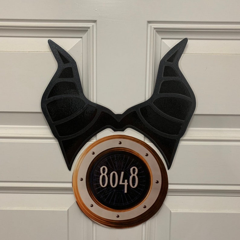 May include: A black and gold door sign with a pair of black horns and a round, gold-rimmed circle with the number 8048 in the center.