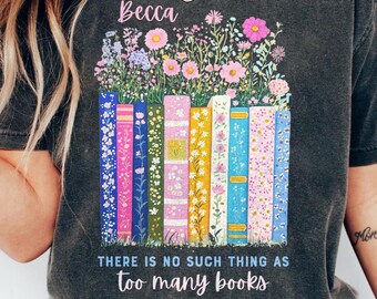 Op de afbeelding: Een donkergrijs T-shirt met een afbeelding van een stapel kleurrijke boeken met bloemen die eruit groeien. De tekst "Becca" is boven de afbeelding gedrukt en de tekst "THERE IS NO SUCH THING AS TOO MANY BOOKS" is onder de afbeelding gedrukt.