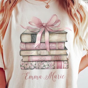 May include: A cream colored t-shirt with a stack of books tied with a pink ribbon and the name "Emma Marie" printed on the front.