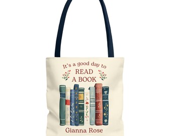 May include: Cream tote bag with navy blue handles. The bag displays a colourful illustration of stacked books and the text "It's a good day to READ A BOOK" with the name "Gianna Rose" beneath the books.