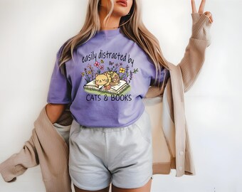 May include: Purple t-shirt featuring a graphic of a cat sleeping on a book with flowers and the text "easily distracted by cats & books".