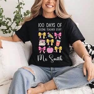Puede incluir: Camiseta negra con un diseño gráfico de lazos rosas y amarillos, libros, lápices y una taza de café. El texto "100 Days of Doing Teacher Stuff" y "Ms. Smith" están impresos en la camiseta.