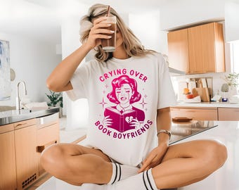May include: White t-shirt with a pink graphic of a crying woman reading a book, with the text "Crying Over Book Boyfriends." The woman is holding a drink with a straw. The shirt is worn by a person sitting on a countertop.