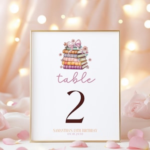 May include: A white table number card with a gold frame. The card features a stack of colorful books with a pink bow and flowers. The text "table 2" is in brown and pink. The bottom of the card reads "Samantha's 13th Birthday 09.18.2030".