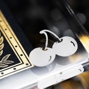 May include: A close-up of a deck of playing cards with a gold border and a black background. The deck is in a clear acrylic case with a white cherry design on the top.