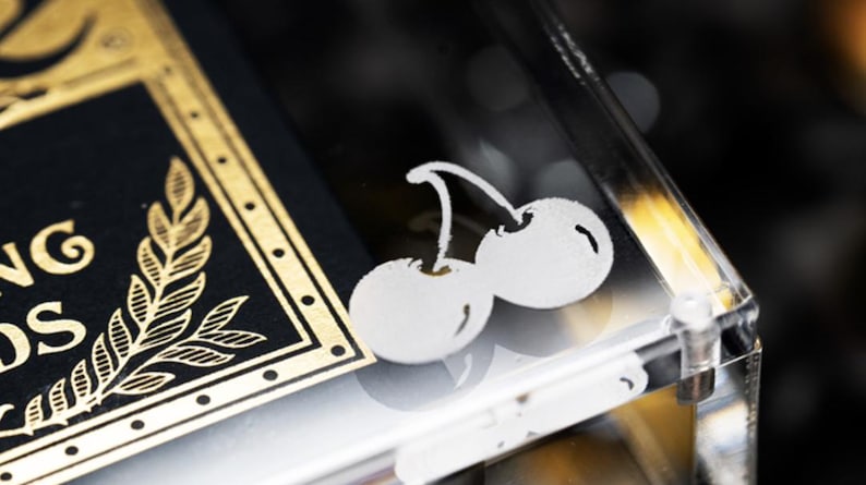 May include: A close-up of a deck of playing cards with a gold border and a black background. The deck is in a clear acrylic case with a white cherry design on the top.