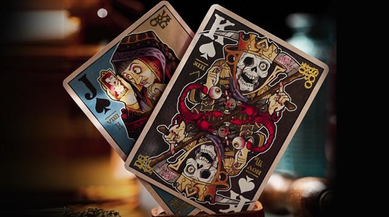 May include: Two playing cards, a Jack of Spades and a King of Spades, from a deck of playing cards with a dark, gothic, and macabre design. The Jack of Spades features a blue background with a woman in a blue and red dress holding a smaller card. The King of Spades features a black background with a skeletal figure wearing a crown and holding a scythe.