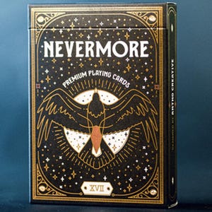 May include: A black and gold playing card box with the word "NEVERMORE" in white. The box features a stylized raven design with the text "PREMIUM PLAYING CARDS" and "XVII". The box has a dark blue background.