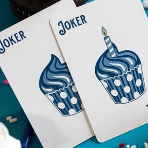 May include: Two playing cards featuring a cupcake design. The cards are white with a blue cupcake illustration and the word "Joker" at the top. One card has a lit candle on top. The background is blue with cupcakes and sprinkles.