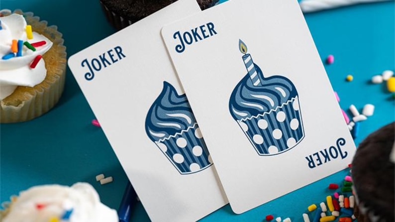May include: Two playing cards featuring a cupcake design. The cards are white with a blue cupcake illustration and the word "Joker" at the top. One card has a lit candle on top. The background is blue with cupcakes and sprinkles.
