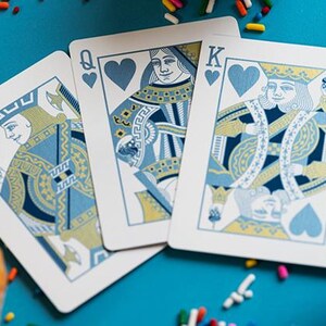 May include: Three playing cards, the Jack, Queen, and King of Hearts, are displayed on a teal surface. The cards feature blue and gold designs. Cupcakes with colourful sprinkles are scattered around the cards.
