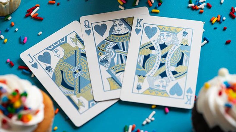 May include: Three playing cards, the Jack, Queen, and King of Hearts, are displayed on a teal surface. The cards feature blue and gold designs. Cupcakes with colourful sprinkles are scattered around the cards.