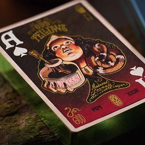 May include: A playing card from the Odd Fellows deck, featuring a woman with a snake wrapped around her arm, holding a jar labeled "Mysteries". The card is the Ace of Spades and has a red background with gold accents.