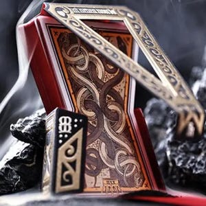 May include: A red and gold playing card holder with a decorative gold frame and a small black card holder. The cards have a brown and gold design with intertwined patterns. The image has a dark, smoky background.