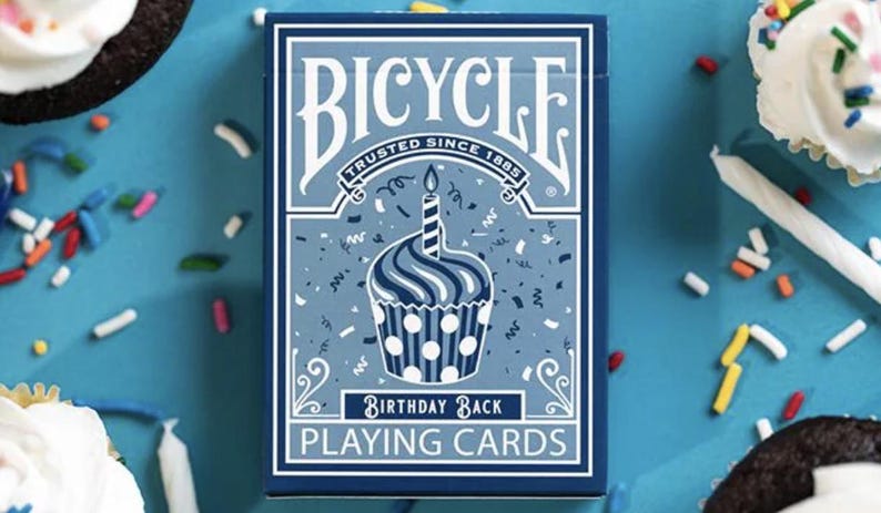 May include: A Bicycle Birthday Back playing card deck featuring a blue and white cupcake design. The deck is surrounded by cupcakes, sprinkles, and candles on a blue background. The text "Bicycle" and "Birthday Back Playing Cards" are visible.