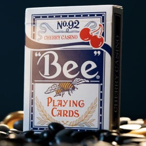 May include: A deck of Bee playing cards with a white background and blue accents. The front of the deck features a bee with a red and yellow striped body and a red cherry with a green stem. The text "No. 92 Cherry Casino" is printed in black at the top of the deck. The text "Bee" is printed in a large, bold font in blue. The text "Playing Cards" is printed in black below the bee.