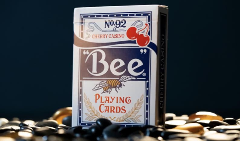 May include: A deck of Bee playing cards with a white background and blue accents. The front of the deck features a bee with a red and yellow striped body and a red cherry with a green stem. The text "No. 92 Cherry Casino" is printed in black at the top of the deck. The text "Bee" is printed in a large, bold font in blue. The text "Playing Cards" is printed in black below the bee.