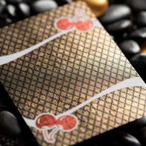 May include: A gold playing card with a black and white checkered pattern and two red cherries on the front. The card is lying on a bed of black stones.
