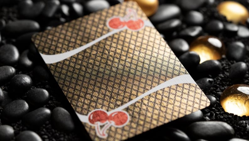 May include: A gold playing card with a black and white checkered pattern and two red cherries on the front. The card is lying on a bed of black stones.