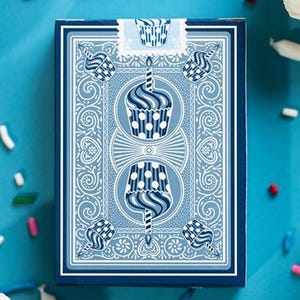 May include: A deck of playing cards with a blue cupcake design. The card box is blue with a white cupcake illustration. Cupcakes and sprinkles are scattered around the cards on a blue surface. A birthday theme.