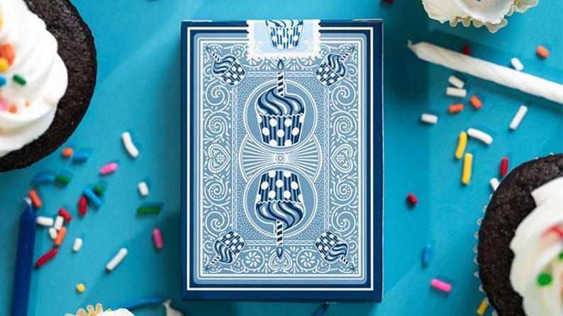 May include: A deck of playing cards with a blue cupcake design. The card box is blue with a white cupcake illustration. Cupcakes and sprinkles are scattered around the cards on a blue surface. A birthday theme.