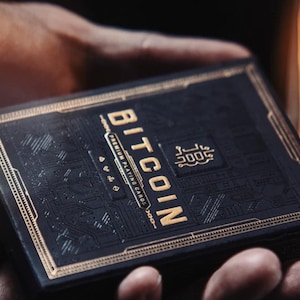 May include: A black deck of playing cards with gold accents. The box features the text "BITCOIN" and "PREMIUM PLAYING CARDS".