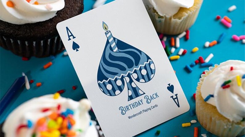 May include: A playing card with a birthday cake design on the Ace of Spades. The card reads "Birthday Back" and is set amongst cupcakes with white icing and colourful sprinkles on a blue surface.