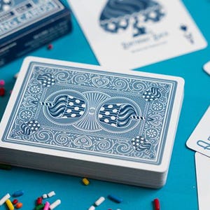 May include: A deck of blue playing cards with a decorative back design, alongside scattered playing cards and a box. The cards feature a detailed pattern and are set against a bright blue surface. A cupcake with sprinkles is in the foreground.