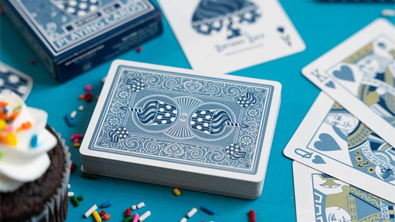 May include: A deck of blue playing cards with a decorative back design, alongside scattered playing cards and a box. The cards feature a detailed pattern and are set against a bright blue surface. A cupcake with sprinkles is in the foreground.