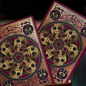 May include: Two playing cards with a dark red background and gold accents. The cards feature a circular design with a central mandala and a border of stylized eyes. The text "The Odd Fellows" is written in a gold script font.