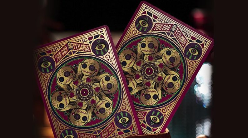 May include: Two playing cards with a dark red background and gold accents. The cards feature a circular design with a central mandala and a border of stylized eyes. The text "The Odd Fellows" is written in a gold script font.