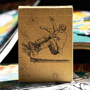 May include: A brown playing card box featuring a black and white illustration of a skateboarder mid-air. The skater is wearing a hat and sneakers. The box has a textured appearance and a small circular logo at the bottom.