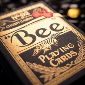 May include: A black box of Bee playing cards with gold foil accents. The box features the text "Bee Playing Cards" and a stylized bee illustration. The box also has a red cherry logo with the text "No. 92 Cherry Casino".