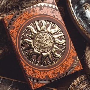May include: A deck of playing cards with a brown and orange design featuring a gold sun with a face in the center. The sun is surrounded by a circular pattern of geometric shapes.