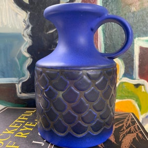 May include: A blue ceramic vase with a textured, scale-like pattern. The vase has a wide mouth and a handle.