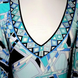 May include: A long-sleeved dress with a V-neckline, featuring a geometric pattern in shades of blue, turquoise, black, and white. The neckline is outlined with a black border and geometric shapes. The dress is displayed on a white mannequin.