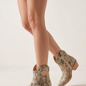 May include: Floral print ankle boots with a pointed toe and a small heel. The boots feature a beige background with pink, yellow, and green floral patterns. The boots have a zipper closure and fringe detailing.