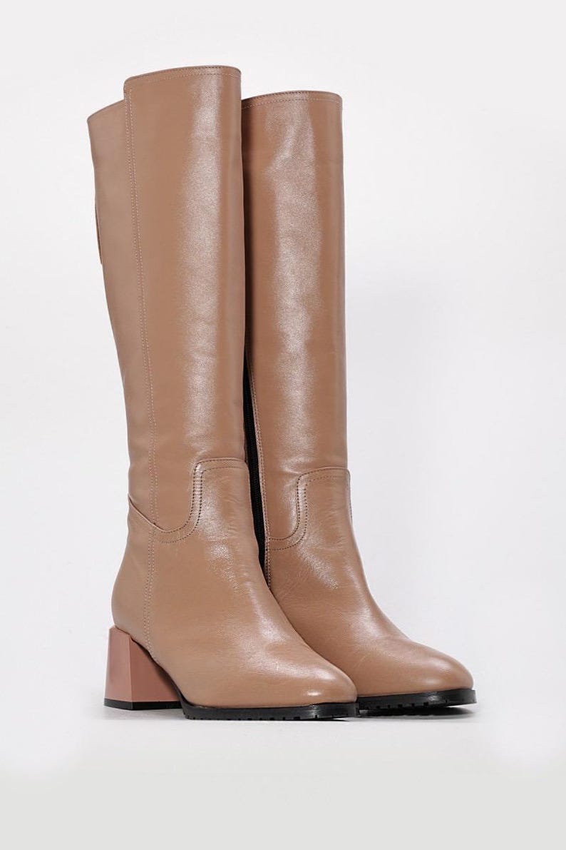 May include: A pair of knee-high boots in a light brown hue. The boots have a square toe and a block heel with a contrasting pinkish-brown color. The soles are black.