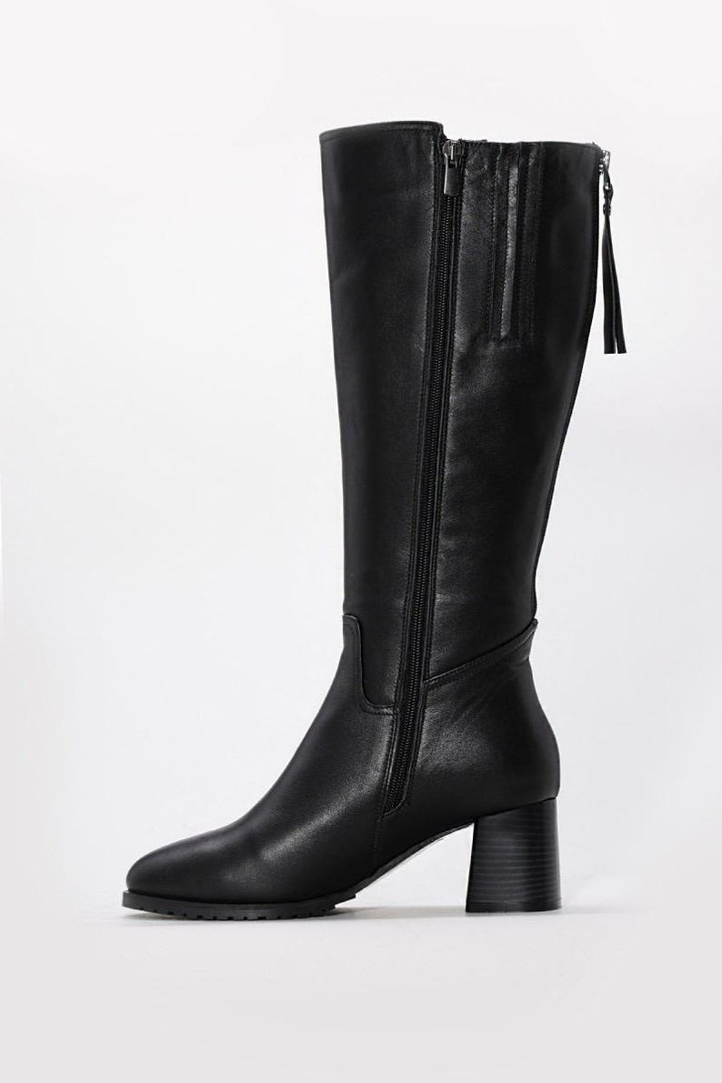 May include: Black knee-high boot with a block heel. The boot features a side zipper and a small tassel detail at the top. The leather boot has a pointed toe and a stacked heel.