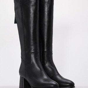 May include: A pair of black knee-high boots. The boots have a block heel and a pointed toe. A zipper and tassel detail are visible on the back of the boot. The boots are made of a smooth, leather-like material.
