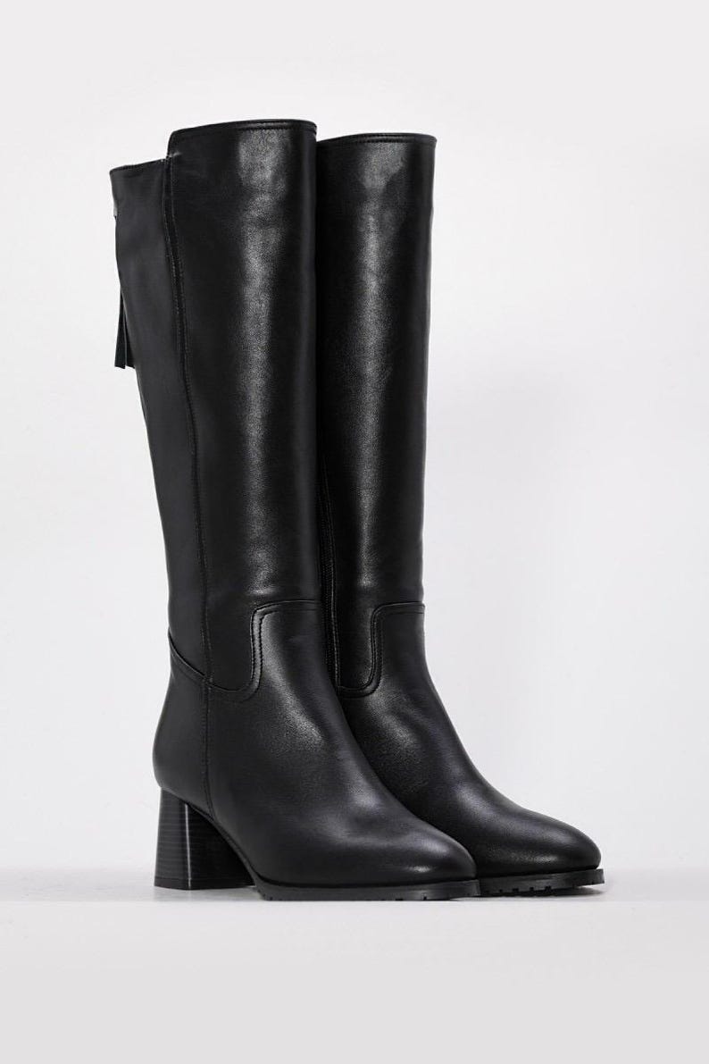 May include: A pair of black knee-high boots. The boots have a block heel and a pointed toe. A zipper and tassel detail are visible on the back of the boot. The boots are made of a smooth, leather-like material.