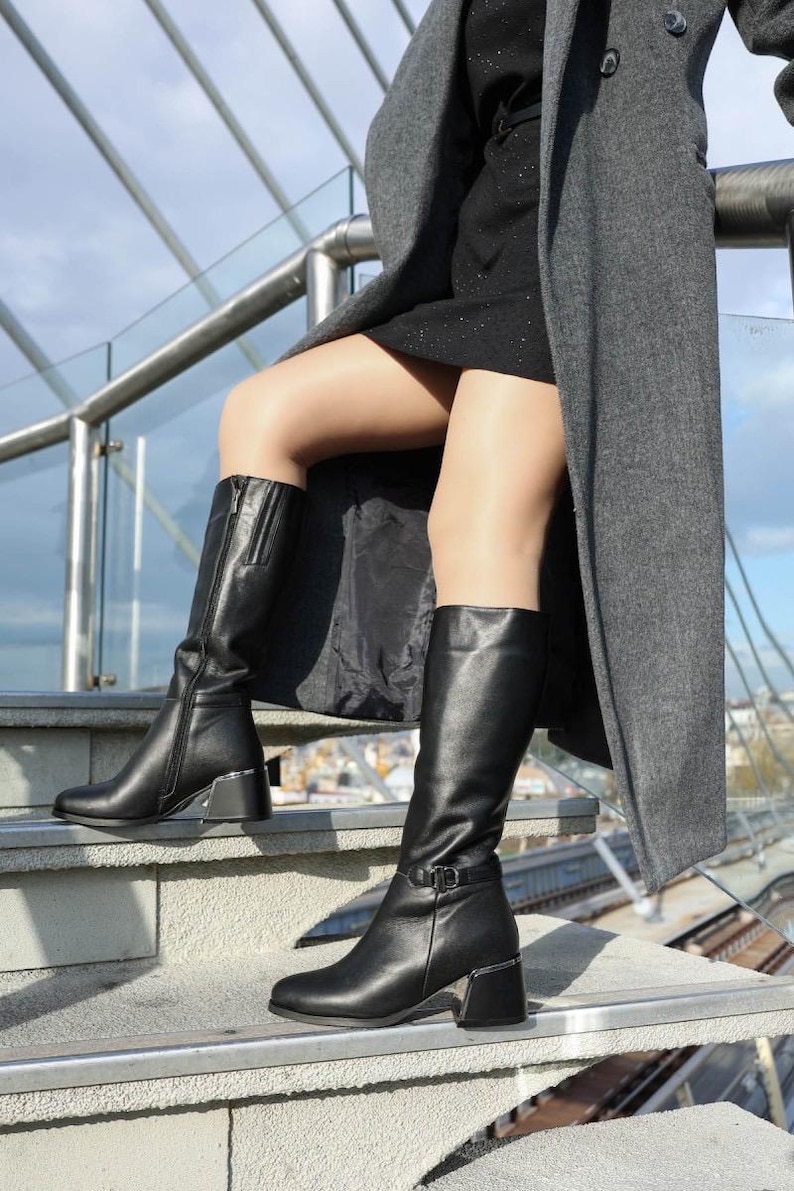 May include: Black leather knee-high boots with silver block heels and zips. One boot has a buckle detail. The boots are worn with a black dress and a grey coat. The boots are on outdoor steps.