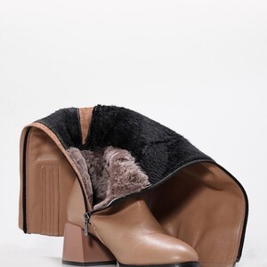 May include: Tan leather knee-high boot with a block heel and a black sole. The boot features a zipper and is lined with faux fur. The top of the boot is folded down, revealing the interior lining. The heel is a matching tan color.
