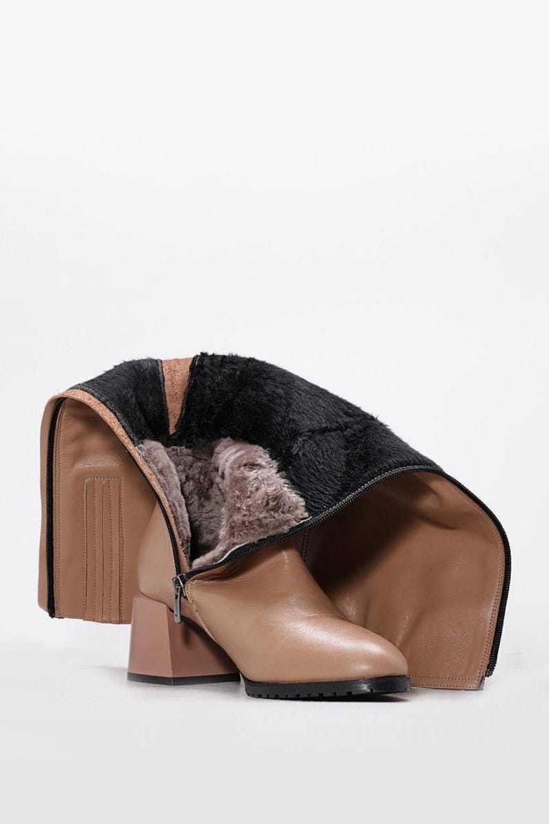 May include: Tan leather knee-high boot with a block heel and a black sole. The boot features a zipper and is lined with faux fur. The top of the boot is folded down, revealing the interior lining. The heel is a matching tan color.