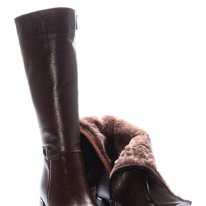 May include: A pair of knee-high brown leather boots with a textured finish. One boot is upright, while the other is partially folded down, revealing a soft, light brown interior lining. The boots have block heels and buckle details.
