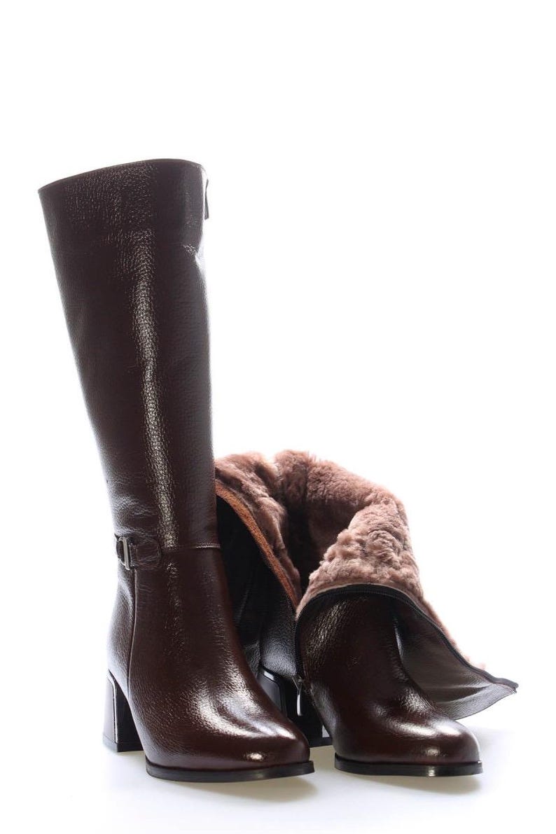 May include: A pair of knee-high brown leather boots with a textured finish. One boot is upright, while the other is partially folded down, revealing a soft, light brown interior lining. The boots have block heels and buckle details.