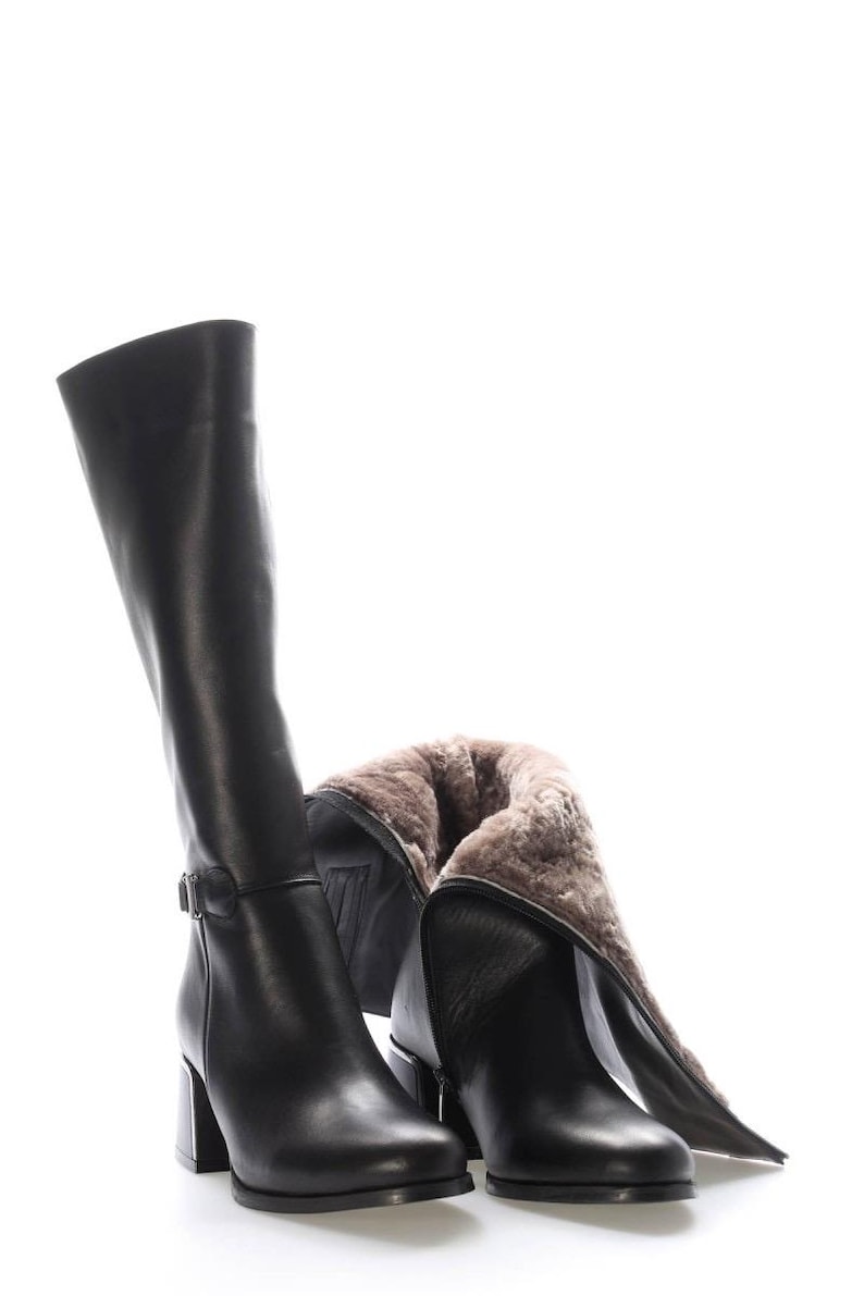 May include: Black leather knee-high boots with a block heel. One boot is upright, the other is partially folded down, revealing a warm, fur-like lining. The boots have a buckle detail near the ankle.