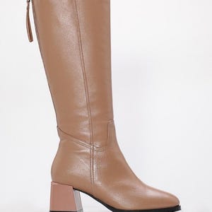 May include: Tan knee-high boots with a block heel. The boots have a zipper on the back and a square toe. The heel is a contrasting light brown color. The boots are made of a smooth leather-like material.