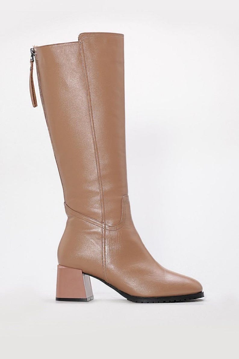 May include: Tan knee-high boots with a block heel. The boots have a zipper on the back and a square toe. The heel is a contrasting light brown color. The boots are made of a smooth leather-like material.