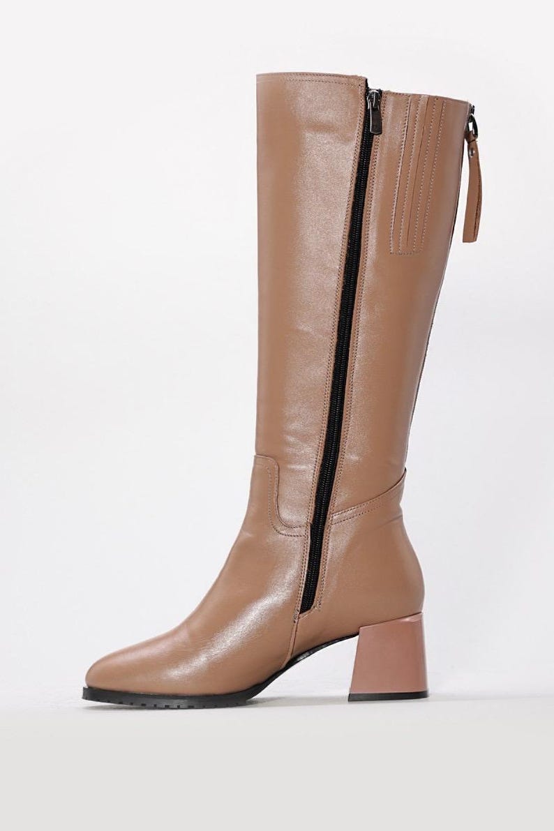 May include: Tan knee-high boot with a black zipper and a block heel. The boot features a rounded toe and a pull tab at the top. The heel is approximately 3 inches high.
